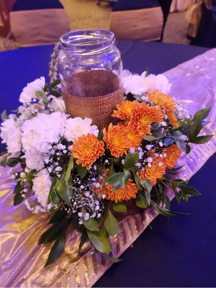 A centerpiece with orange and white chrysanthemums surrounding a glass jar. The arrangement sits on a gold and purple table runner, adding a pop of color to the table setting.