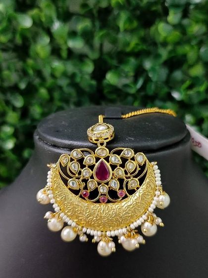 A beautiful chaand bali style maang tikka with a textured gold finish, kundan work, and a central ruby-colored stone.