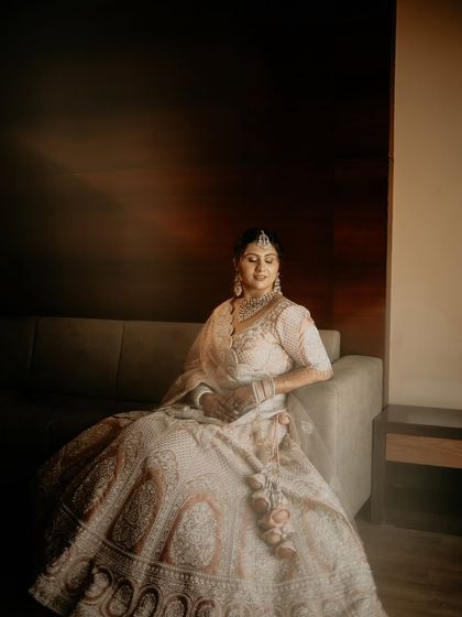 A full-length bridal portrait in a quiet, contemplative setting. The bride sits gracefully on the sofa, her magnificent lehenga spread around her, looking like royalty.