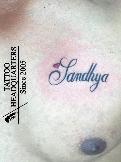 The name "Sandhya" with a small red heart over it, tattooed on the chest. A classic and loving tribute.