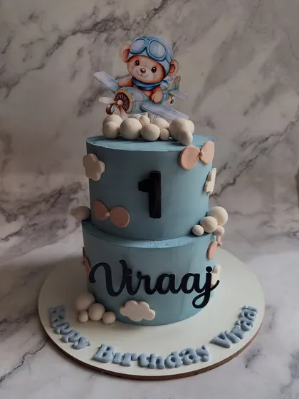 Taking flight for a first birthday! This custom two-tier cake features a hand-modeled pilot teddy bear, fluffy clouds, and personalized details to make a little one's milestone celebration truly special.