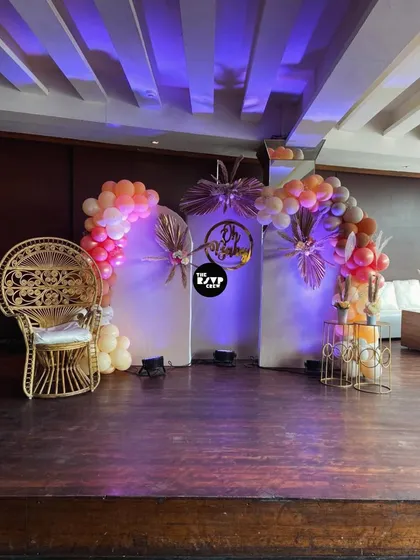 A chic and modern stage design for a baby shower. We used clean white panels, dramatic purple uplighting, and metallic palm accents to frame the "Oh Baby" sign. The iconic peacock throne and asymmetrical balloon arches complete this stylish setup.