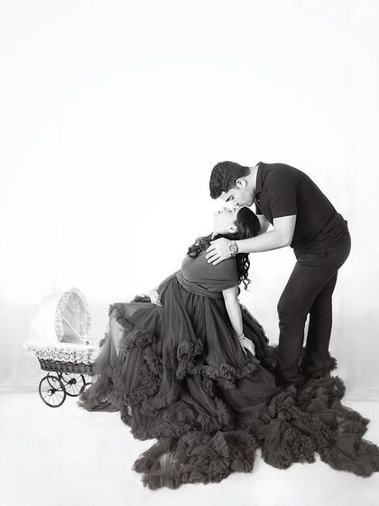 A dramatic and artistic black and white photo. The dad-to-be leans in to kiss his partner's forehead as she reclines in a stunning dark ruffled gown.