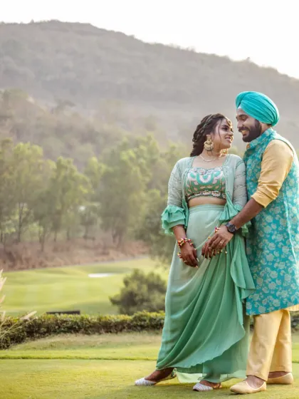A Sikh couple shares a loving gaze amidst a lush green landscape with hills in the background. Their vibrant traditional attire provides a beautiful contrast to the natural setting, blending royalty with nature.