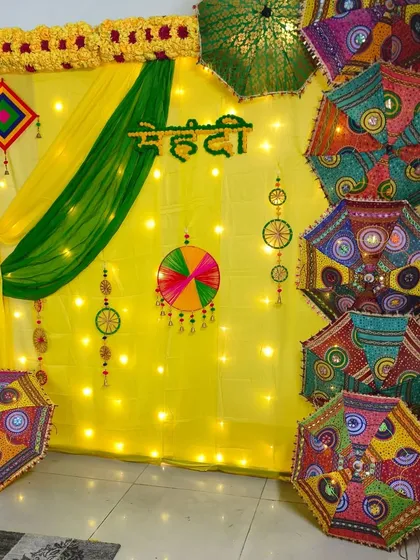 For a traditional Mehendi ceremony, I create a vibrant backdrop using yellow and green fabric drapes, warm fairy lights, and a collection of colorful Rajasthani umbrellas with mirror work. The Hindi "Mehendi" lettering adds an authentic touch.