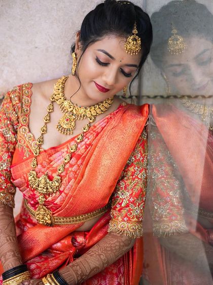 A stunning bridal portrait in a vibrant orange silk saree. This look features classic winged eyeliner and a bold red lip, paired with heavy temple jewellery for a truly traditional South Indian wedding style.