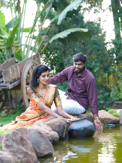 A serene pre-wedding photo by a small pond. The couple is interacting with the water, creating a natural and romantic scene in their traditional outfits.