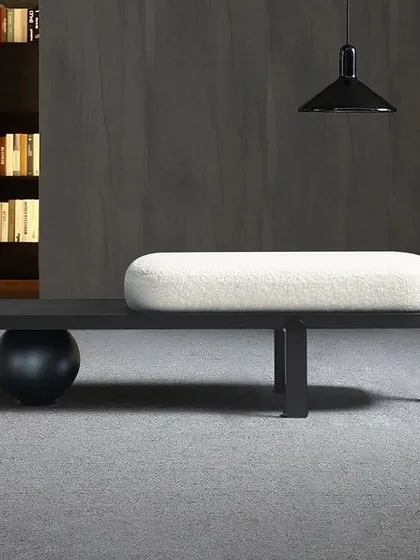 This modern bench features a white Bouclé upholstered cushion on an asymmetrical black base, with one end supported by a metal sphere and the other by simple legs.