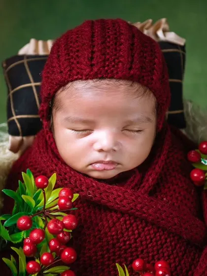 A close-up of a newborn wearing a red knitted hat, surrounded by festive red berries. This is a great example of a seasonal or holiday-themed photoshoot.