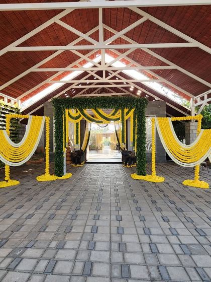 A traditional wedding entrance arch decorated with lush green foliage and garlands of yellow and white flowers, creating a warm and auspicious welcome for guests.
