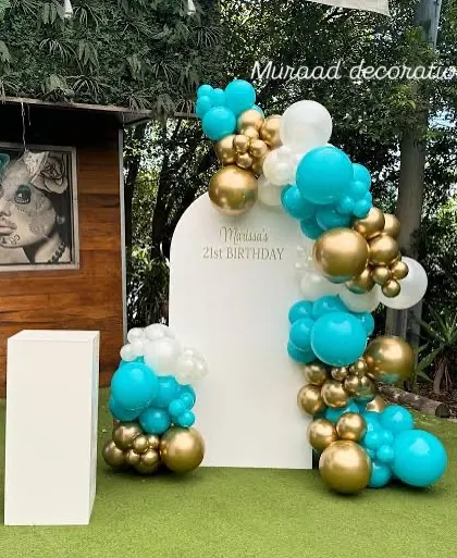 A simple and elegant outdoor backdrop for a 21st birthday. The white arched panel is decorated with a beautiful balloon garland in turquoise, white, and gold, which stands out against the green lawn.