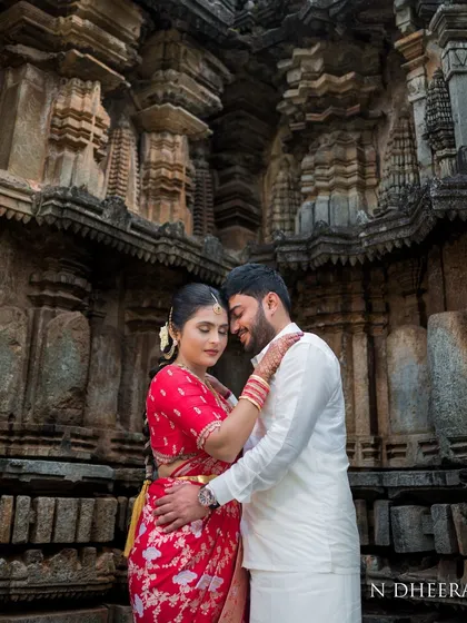 A romantic couple shot featuring the bride in her red saree and custom blouse. The traditional setting enhances the timeless quality of her outfit.