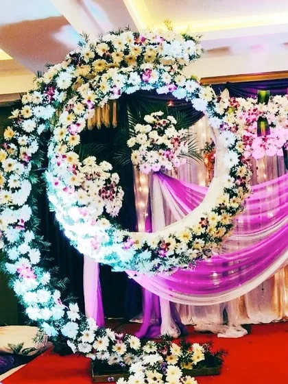 A close-up view of an illuminated floral crescent moon structure. The built-in lights add a magical glow to the white and purple flowers, perfect for an evening ceremony.