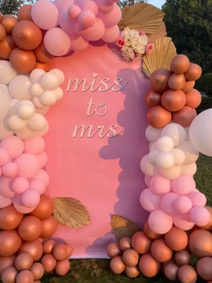 An outdoor bridal shower setup with a 'Miss to Mrs' backdrop. The balloon arch in shades of pink, white, and bronze looks beautiful against the natural green lawn.