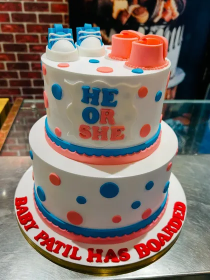 A fun two-tier "He or She" gender reveal cake with blue and pink polka dots and tiny baby shoes on top.