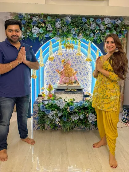 A happy client posing with the 'Aqua Blue' Ganpati decor. This setup features a beautiful floral arch and traditional hanging elements, creating a picture perfect corner for festive family photos.