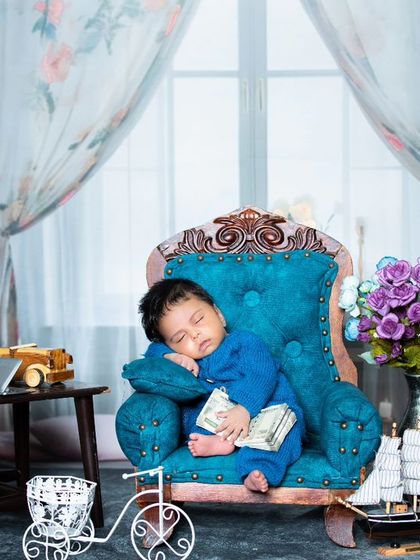This newborn is sleeping like a king on a miniature velvet armchair, holding a tiny book.