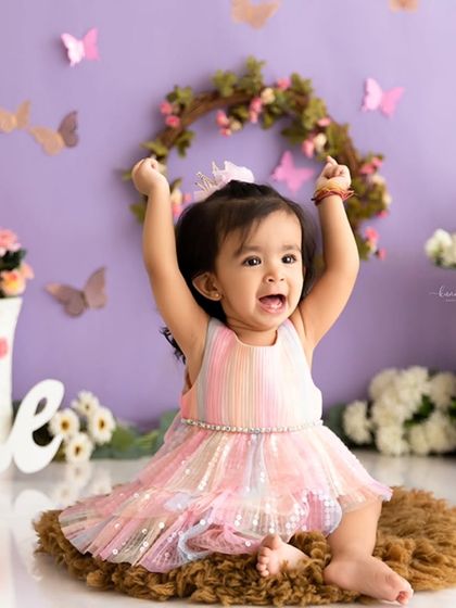 A moment of pure celebration captured before the cake smash, with the birthday girl in a rainbow dress against a lavender butterfly backdrop.