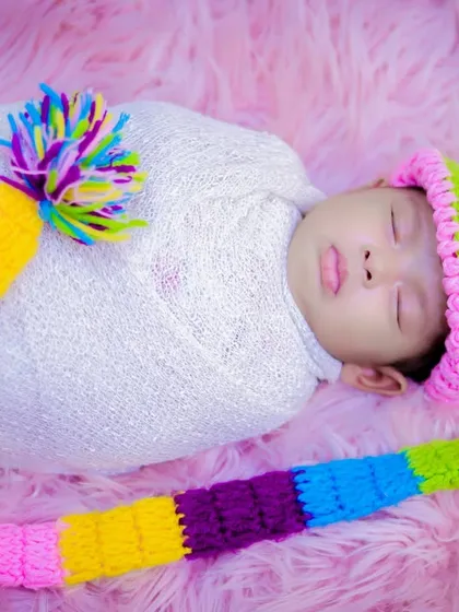 A newborn wrapped in a white swaddle wearing a playful, colorful knitted hat, sleeping soundly on a pink fur background.