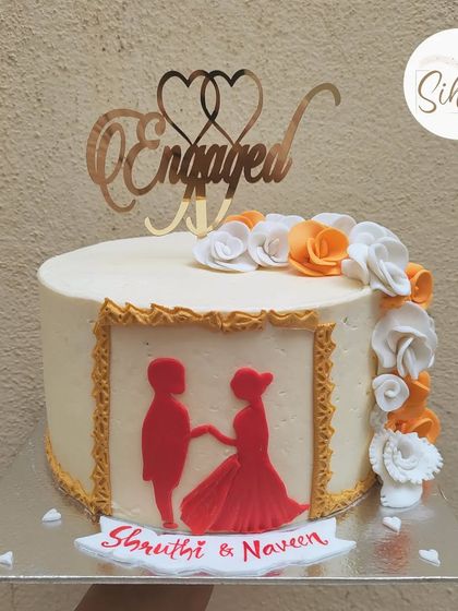 A lovely engagement cake featuring a framed silhouette of a couple in red and orange. The design is completed with hand-piped sugar flowers and a gold acrylic topper.