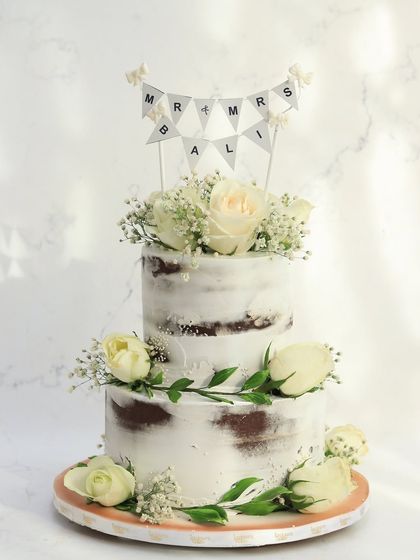 This two-tiered semi-naked cake offers a rustic yet charming look. Adorned with fresh white roses and baby's breath, it's a simple and beautiful centerpiece for a wedding celebration, personalized with a "Mr & Mrs" banner.
