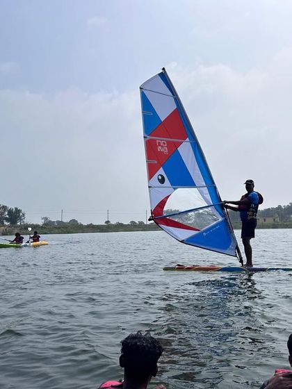 An instructor demonstrates windsurfing technique to a group of students watching from the water in Ambigarahalli.