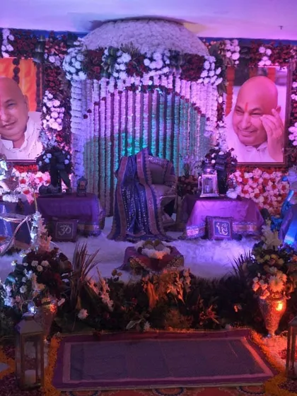 A devotional setup featuring a purple and blue color theme. The gaddi is placed between two portraits of Guru Ji, with the entire stage decorated with flowers and lit by traditional lanterns.