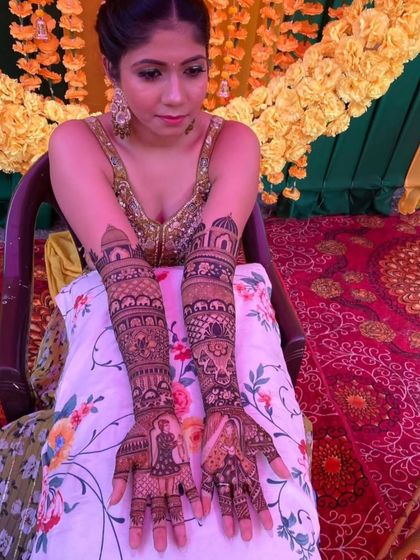 A seated bride proudly displays her full-arm narrative mehendi, featuring bride and groom figures on the palms and palace architecture on the forearms.