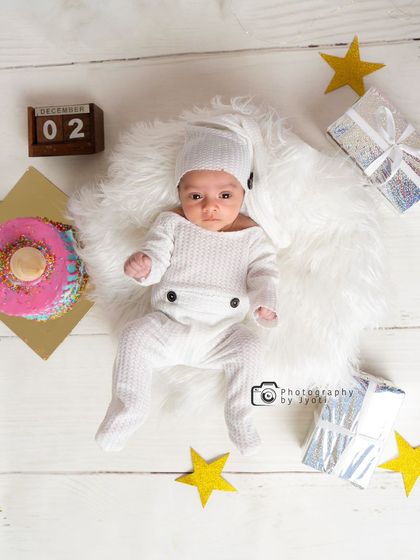 A creative flat-lay photo of a newborn on a soft white rug. The scene is decorated with a prop cake, stars, and a calendar block showing the date, perfect for a birth announcement.