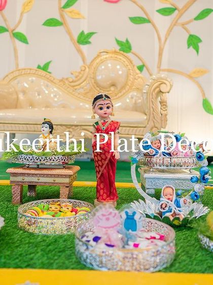 This baby shower platter features a beautiful doll in a traditional saree, surrounded by sweets and gifts. I can customize the dolls' attire to match your event's theme.
