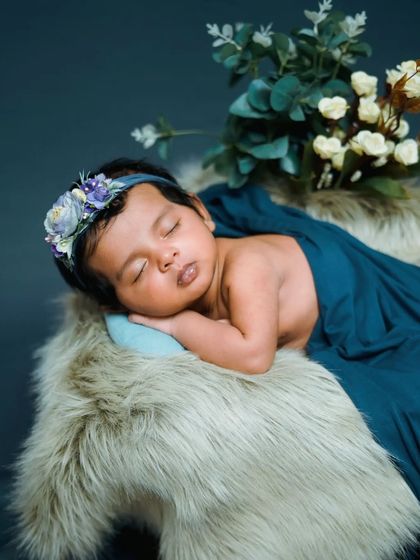 A peaceful newborn pose on a soft fur rug, adorned with a delicate floral headband. The deep teal wrap adds a beautiful pop of color.