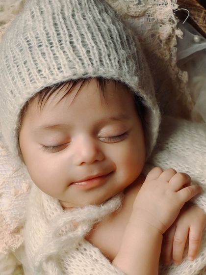 A close-up of a baby's serene, smiling face while wearing a soft, knitted bonnet. The neutral colors and soft textures create a feeling of warmth and peace.