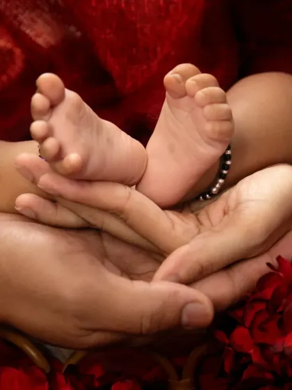 A close-up, symbolic photo of a newborn's tiny feet, complete with a traditional black anklet, held securely and lovingly in their parents' hands.