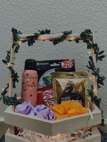 This hexagon-shaped wooden basket is filled with a mix of fun and practical items. It includes a deodorant, a face mask, colorful scrunchies, and a variety of chocolates.