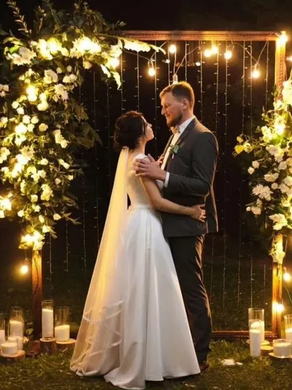 A romantic photo spot for a pre-wedding shoot or reception. The couple is framed by a wooden structure decorated with flowers and fairy lights, perfect for evening pictures.