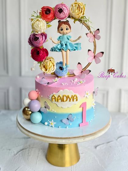 A whimsical first birthday cake for Aadya. This pastel rainbow cake is decorated with butterflies, colorful spheres, and a beautiful doll on a floral swing topper.