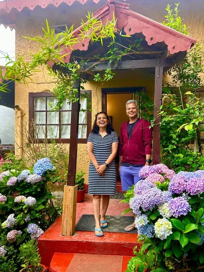 Happy guests stand at the entrance of a cottage at Jungle Lore Birding Lodge, framed by colorful flowers. We pride ourselves on creating a welcoming and memorable experience for everyone who stays with us.