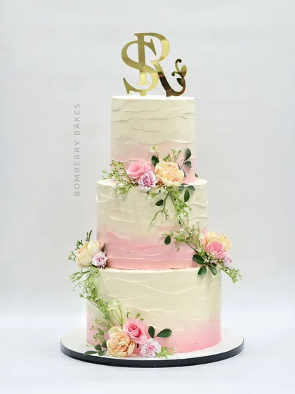 A three-tier buttercream wedding cake with a soft pink ombre effect and adorned with fresh pastel flowers and greenery.