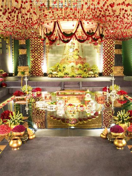 A detailed shot of the main stage and mandap area. The backdrop features a large, beautifully lit painting of a deity, surrounded by a sea of flowers and traditional brass vessels, creating a rich and reverent focal point.