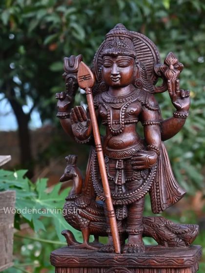 A 24-inch wooden sculpture of Lord Murugan with his peacock, finished in a rich wax brown and placed in a garden setting.