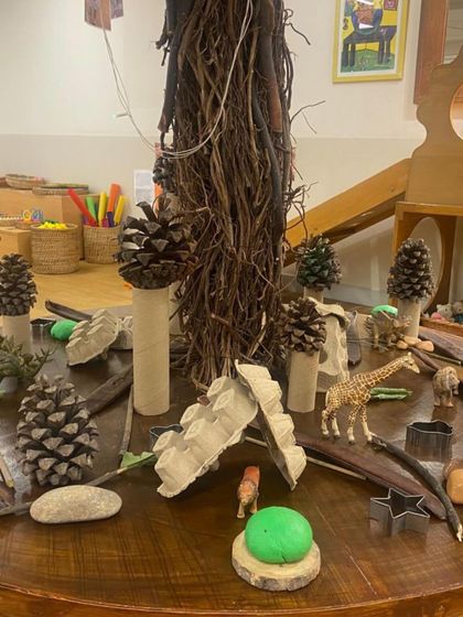 A "loose parts" invitation centered around a tree-like structure made of twigs. Pinecones, wooden blocks, and animal figures are provided, encouraging children to build a forest ecosystem and tell stories about the animals that live there.