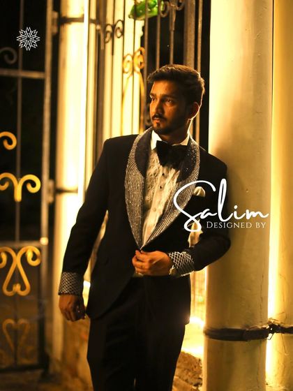 A model leaning against a pillar, wearing a black tuxedo with crystal-studded lapels and cuffs. The professional shot highlights the elegance of the design.