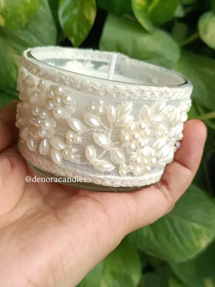 A hand-held view of a jar candle wrapped in white floral lace with pearl details.