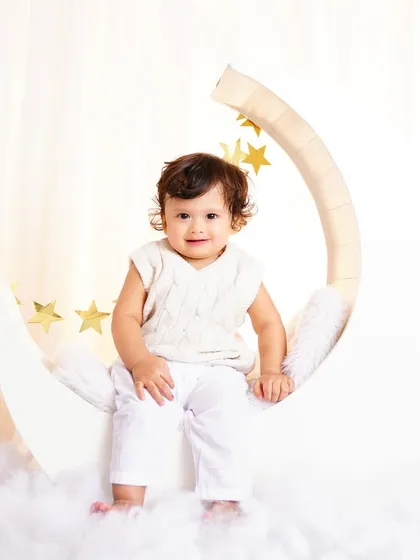 A smiling baby sitting on a white crescent moon prop, dressed in an adorable all-white outfit. A heavenly and sweet portrait.