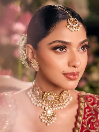 A detailed close-up of our signature red bridal look. You can see the flawless, soft-focus skin, the gentle bronze tones on the eyes, and the classic floral bun that completes this picture of elegance.