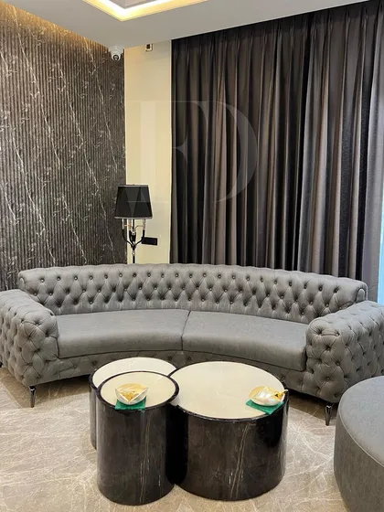 This living room features a curved, tufted Chesterfield sofa in grey, paired with a matching circular ottoman. The set creates a cohesive and elegant look, grounded by dark marble nesting tables.