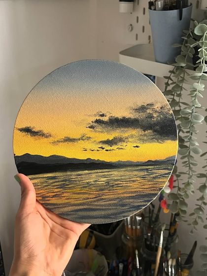 My finished round canvas painting of a yellow sunset, held up in my studio. The light catches the details of the water's reflection beautifully.