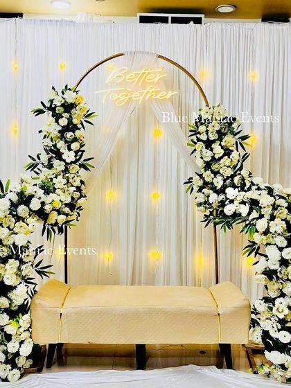 A classic Roka setup featuring a golden circular arch adorned with white flowers. The 'Better Together' neon sign and fairy light backdrop create a warm and inviting ambiance for any engagement or anniversary celebration.