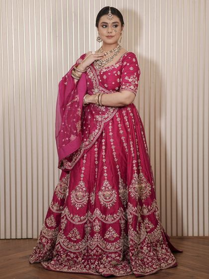 A full view of a bride in a deep magenta lehenga. The outfit is paired with a matching Kundan necklace set, showing a complete and cohesive look available for rent.