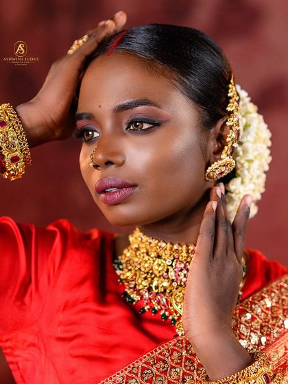 This portrait showcases a beautiful South Indian bridal look, with jasmine flowers in the hair and traditional gold jewelry. The warm, reddish backdrop complements the bride's red saree perfectly.